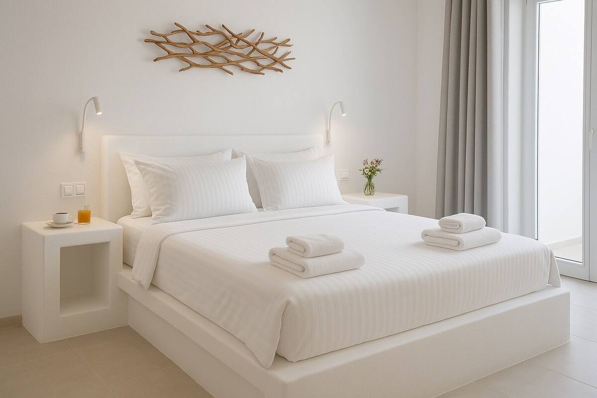 Quarto de hotel moderno com uma cama grande, roupa de cama branca, um painel decorativo e uma mesa de cabeceira com uma xícara e um copo.