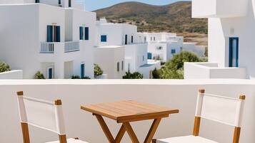A terrace with two wooden chairs and a table, overlooking a white building complex with blue accents and a view of a hill.