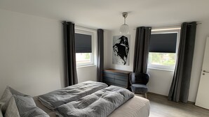 A modern bedroom with a bed, a chair, a desk, and a large window with curtains.