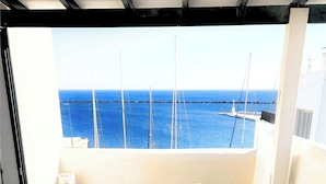 Una mesa de madera con cojines blancos y una planta en maceta en el centro, ubicada en un balcón con vistas al mar y a barcos.