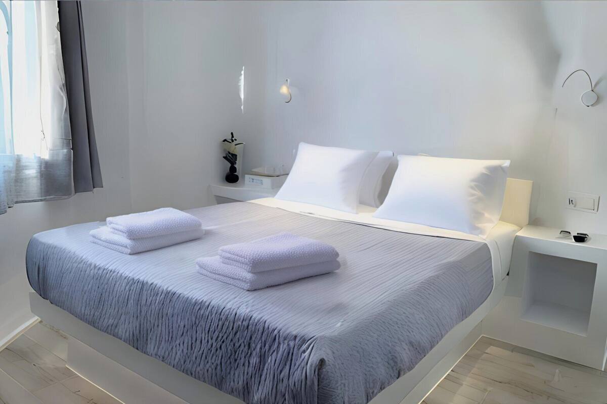 Un dormitorio moderno con una cama grande, ropa de cama blanca, una mesita de noche con lámpara y una planta pequeña.