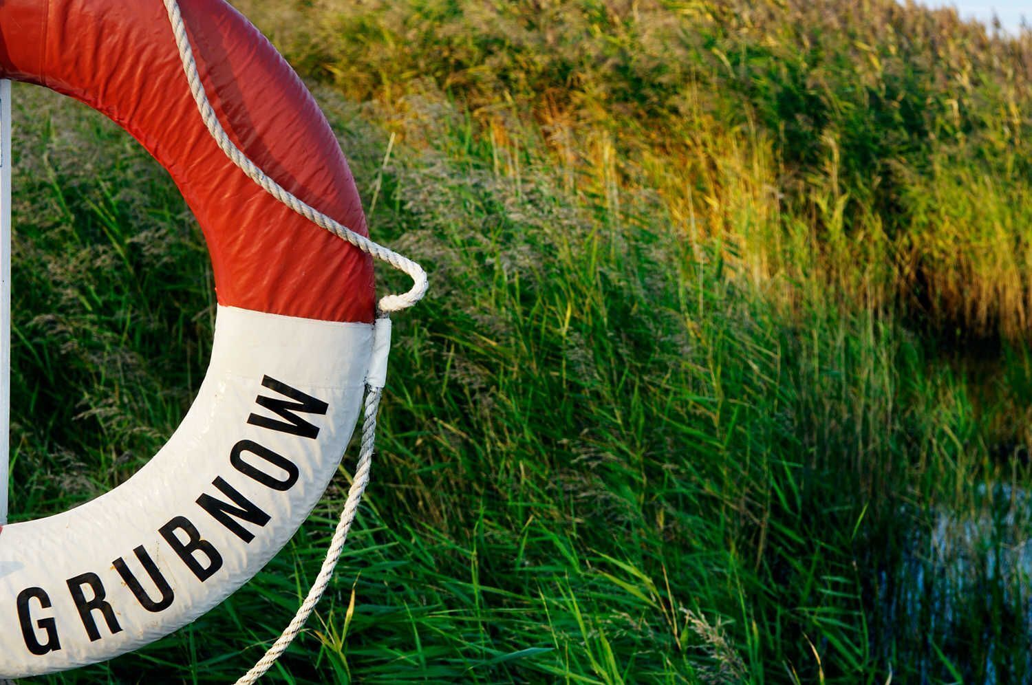 Une bouée de sauvetage portant l’inscription « GRUBNOW », attachée à une corde, le tout sur un fond d’herbe.