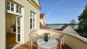 A balcony with a wooden table and chairs, a vase of flowers, and a view of a lake and buildings. - Apartment 7 with lake view and large balcony - Gut Grubnow Rügen (Neuenkirchen)