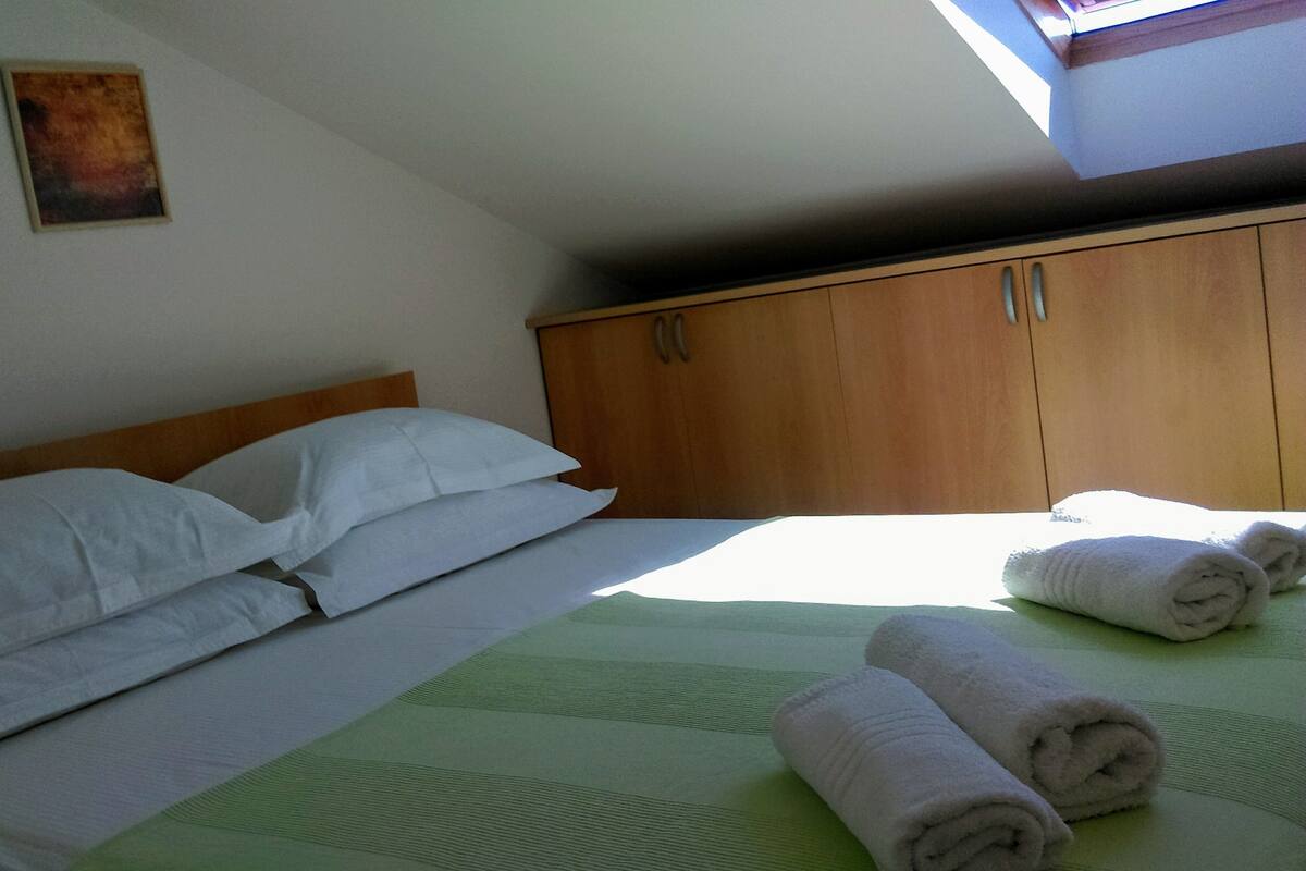 A bedroom with a bed, pillows, a green bedspread, and rolled towels on the floor.