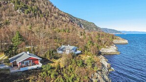 Une cabane au bord de la mer, avec une façade rouge et un balcon, entourée d’une forêt dense et d’une côte rocheuse.