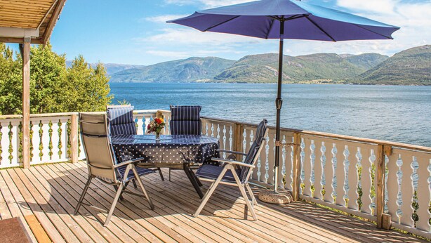 Une terrasse en bois avec une table et des chaises, un parasol violet et une vue sur un lac, avec des montagnes en arrière-plan.