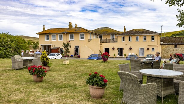 An outdoor seating area with wicker furniture and potted plants, a building with a balcony, and a clear sky. - Haddon House Hotel (Bridport)