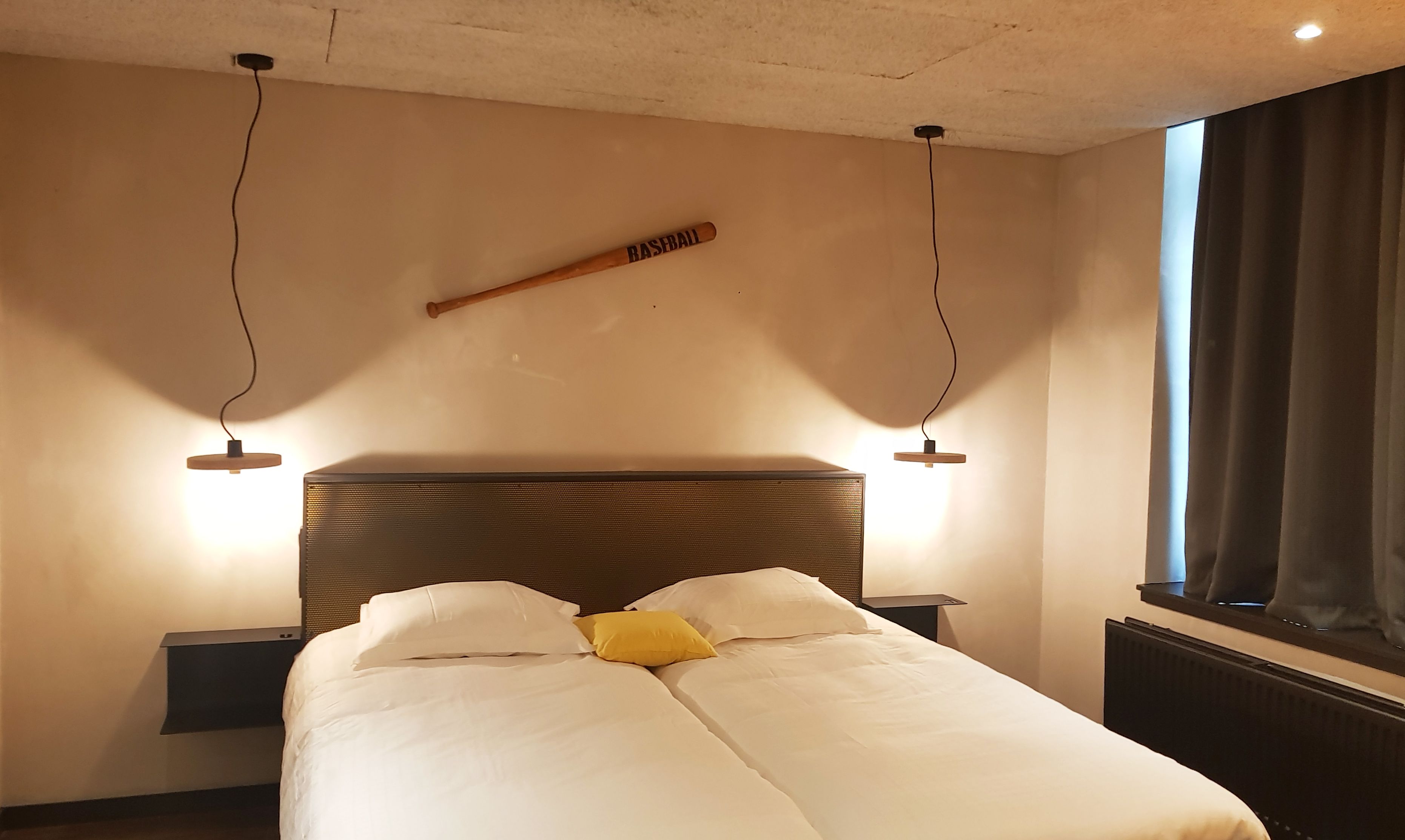 Una cama bien hecha con almohadas blancas y amarillas, un bate de béisbol de madera colgado en la pared y dos lámparas de noche.
