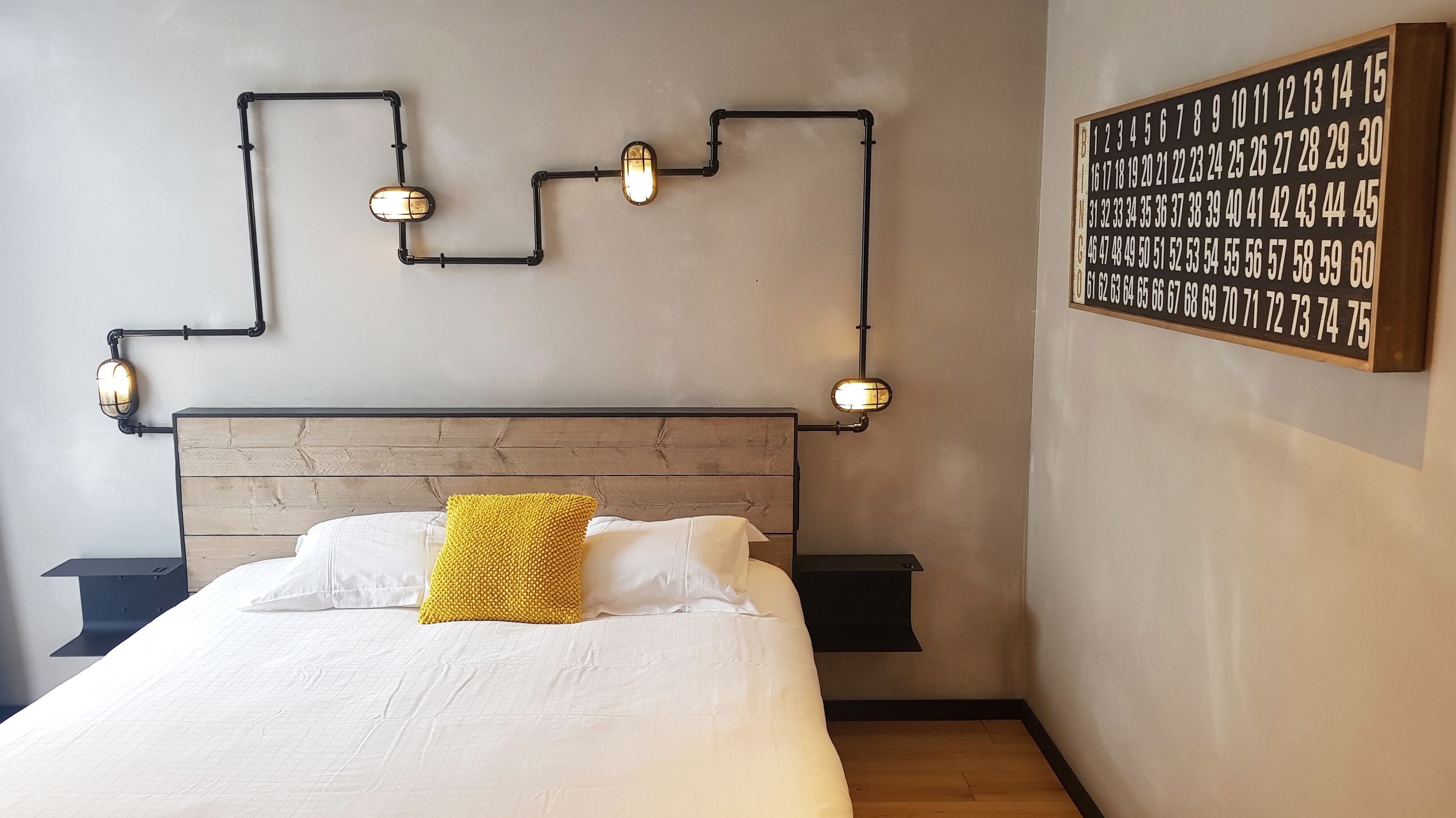 A neatly made bed with white linens and a yellow accent pillow, a headboard with a wooden texture, and a wall-mounted light fixture with a number sequence.