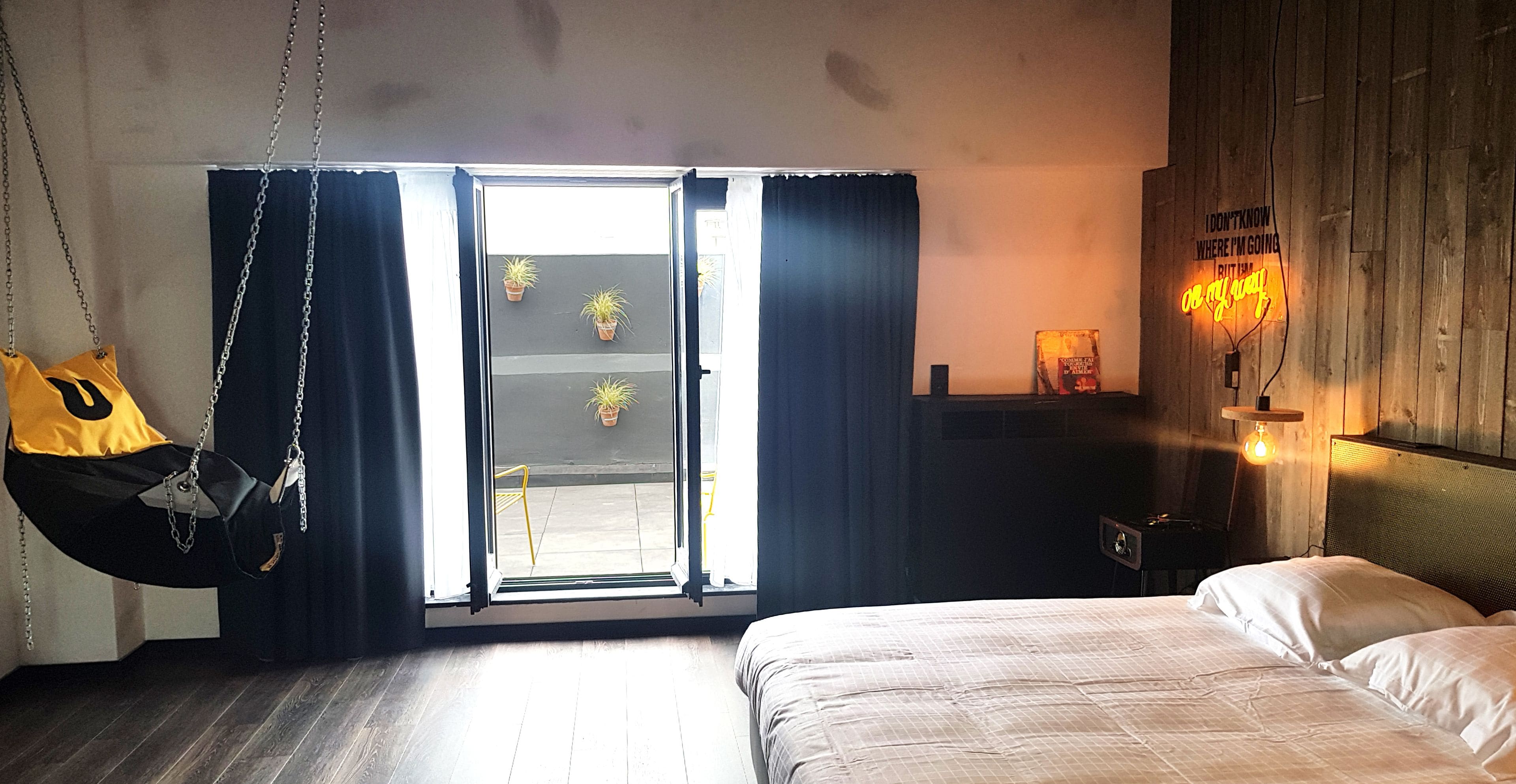 Ein Schlafzimmer mit einem Bett, einem Hängestuhl, einem Fenster mit Vorhängen und einer Neonleuchtreklame.