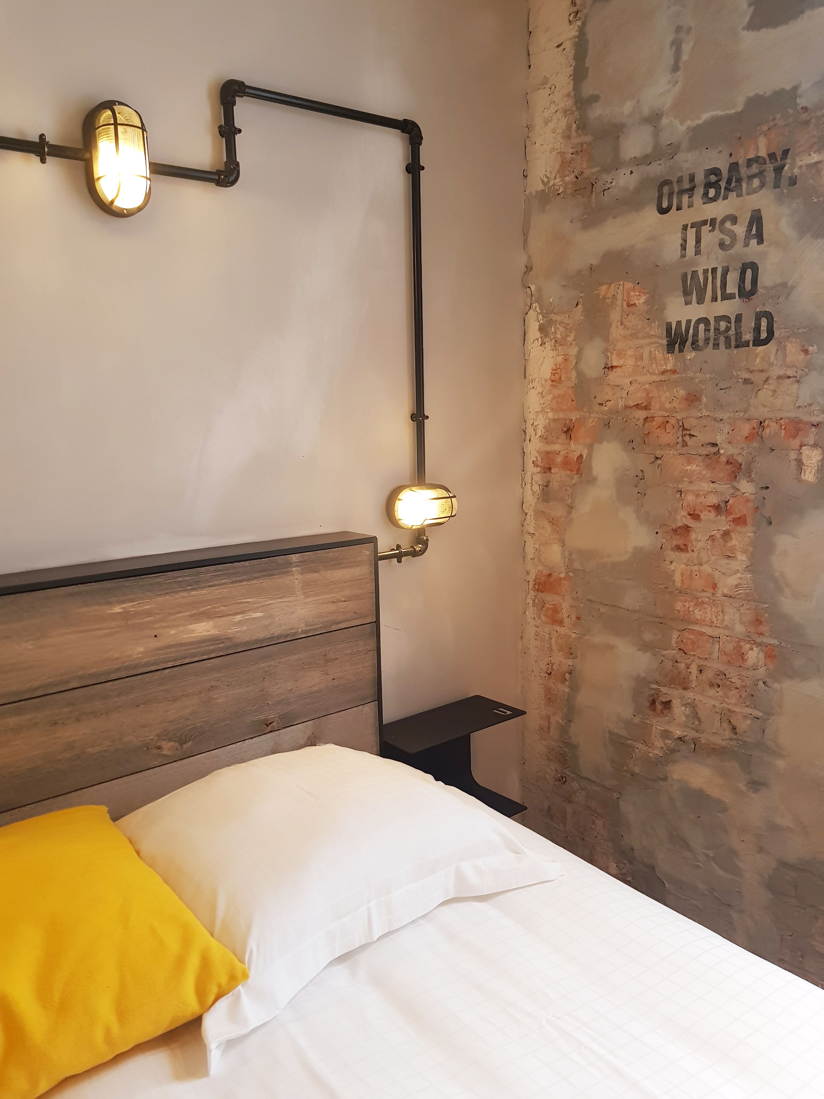 A bedroom with a wooden headboard, a bed with white bedding and a yellow pillow, a nightstand, and a wall with exposed brick and a motivational quote.