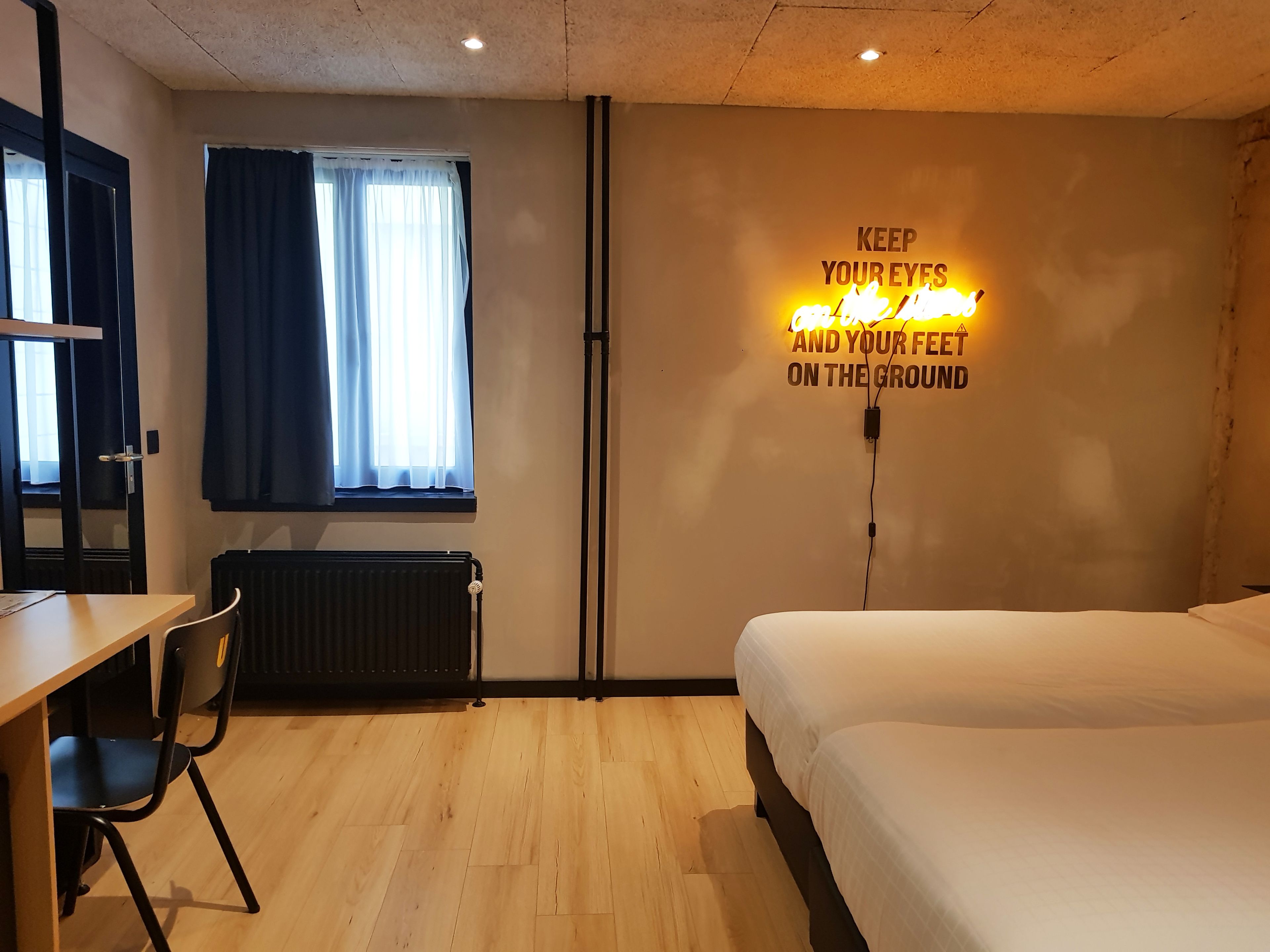 Een slaapkamer met een bed, een bureau, een stoel, een radiator en een muur met een inspirerende quote.