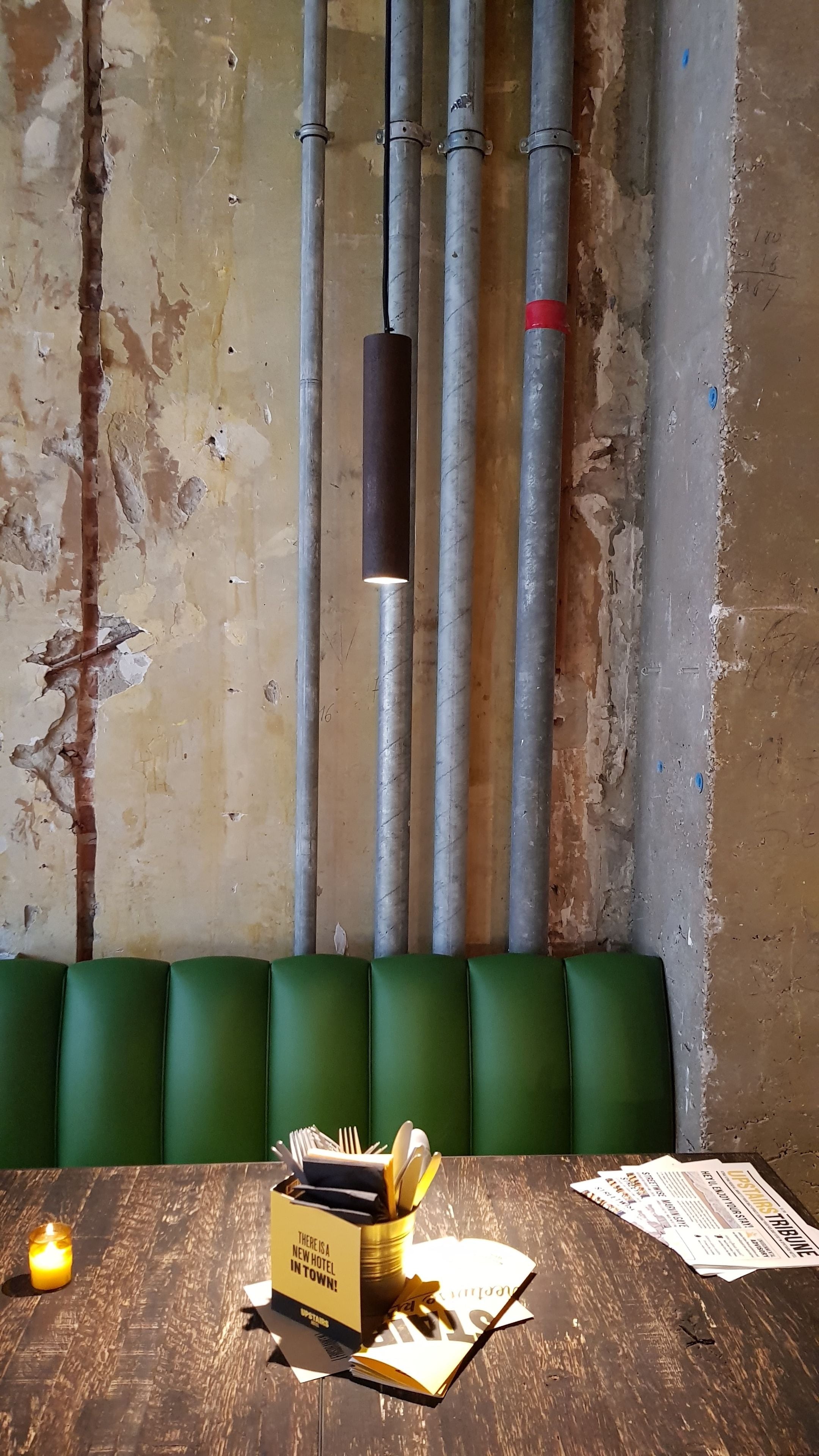 A restaurant interior with exposed pipes, green upholstered booths, and a table with a candle, a newspaper, and a box of utensils.