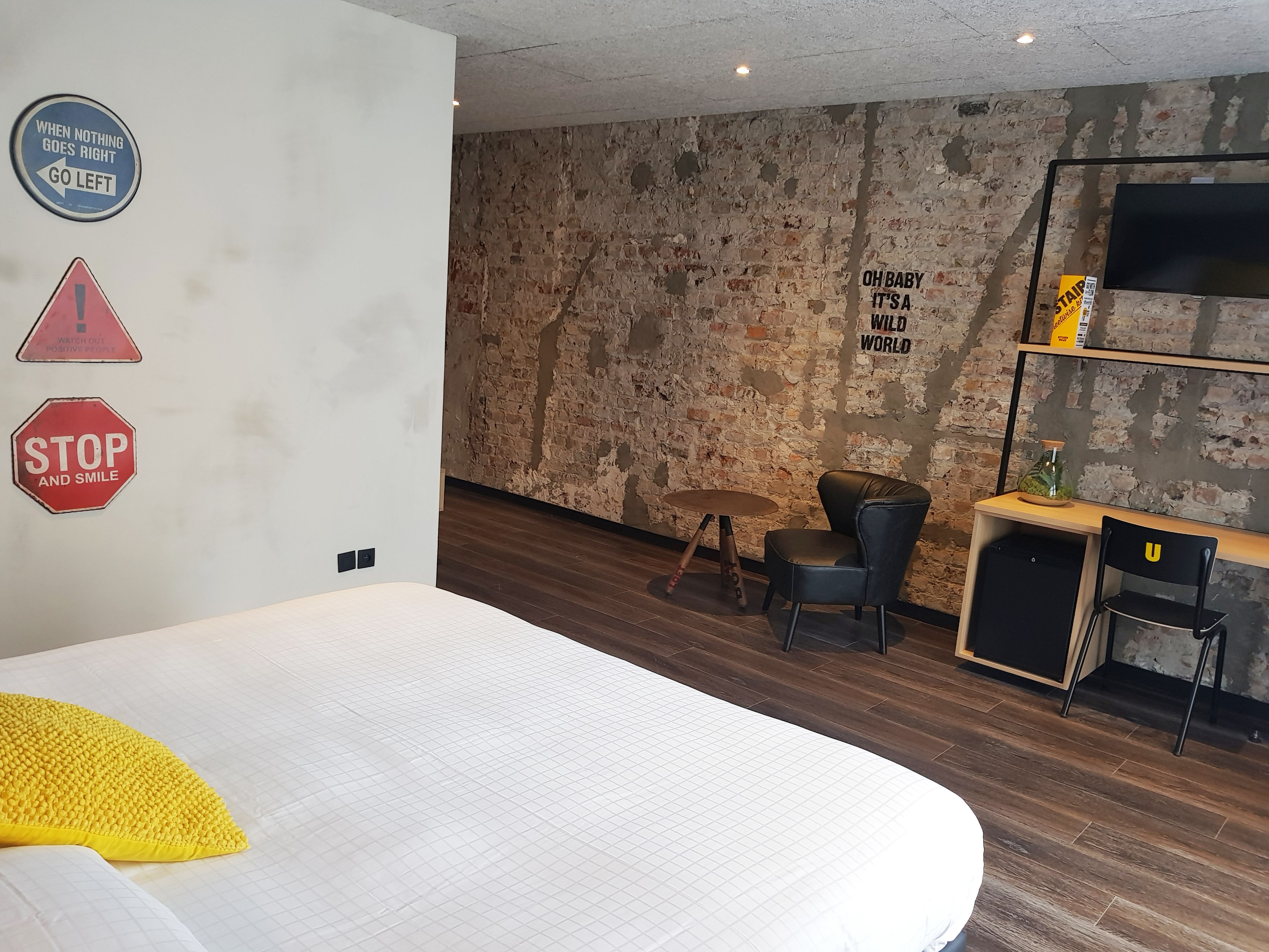 A modern hotel room with a brick wall, a bed with a yellow pillow, a desk with a chair, and motivational wall stickers.