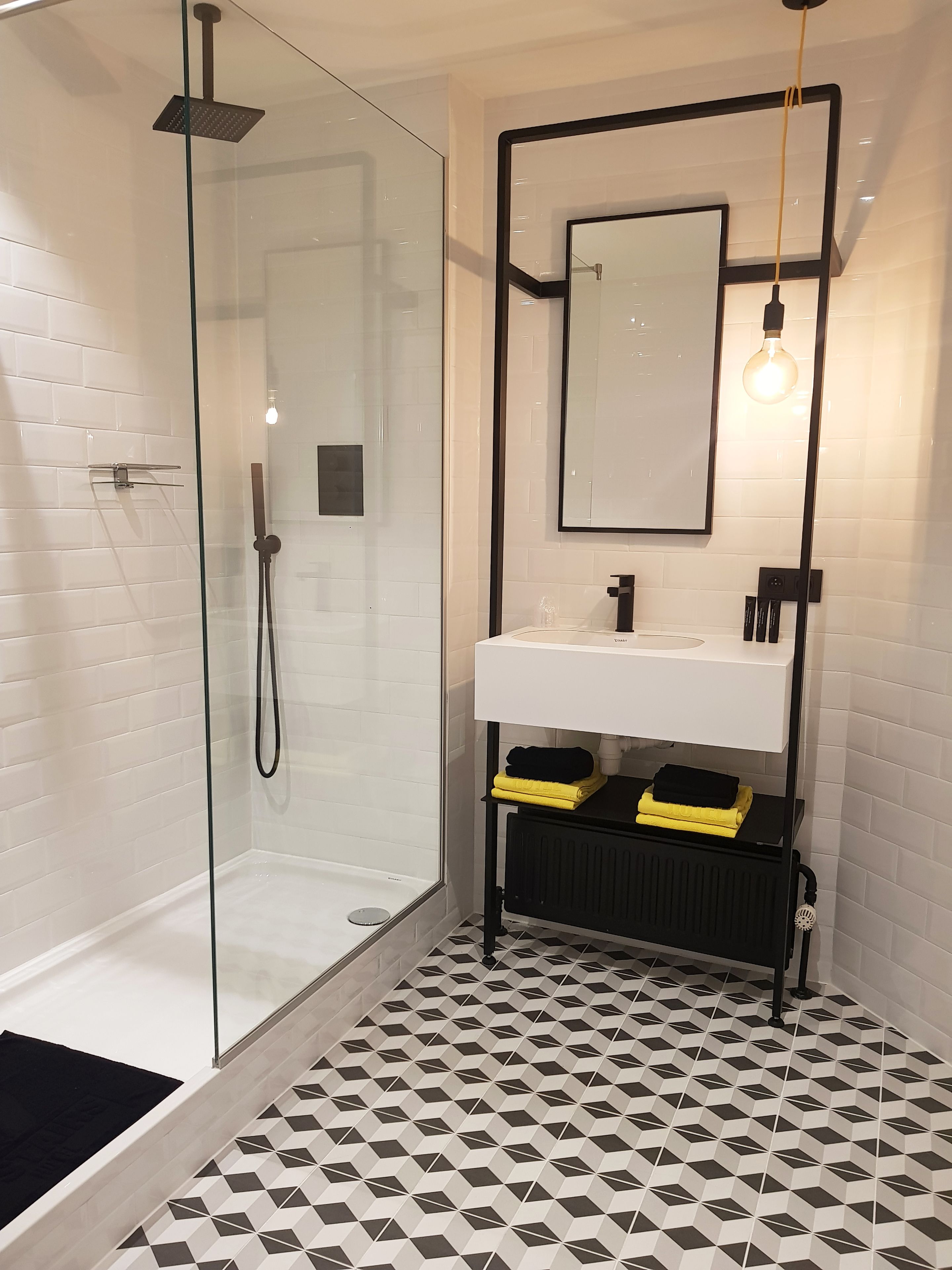Een moderne badkamer met een douchecabine van glas, een wit wastafeltje met een zwart aanrechtblad en een vloer met een geometrisch patroon.