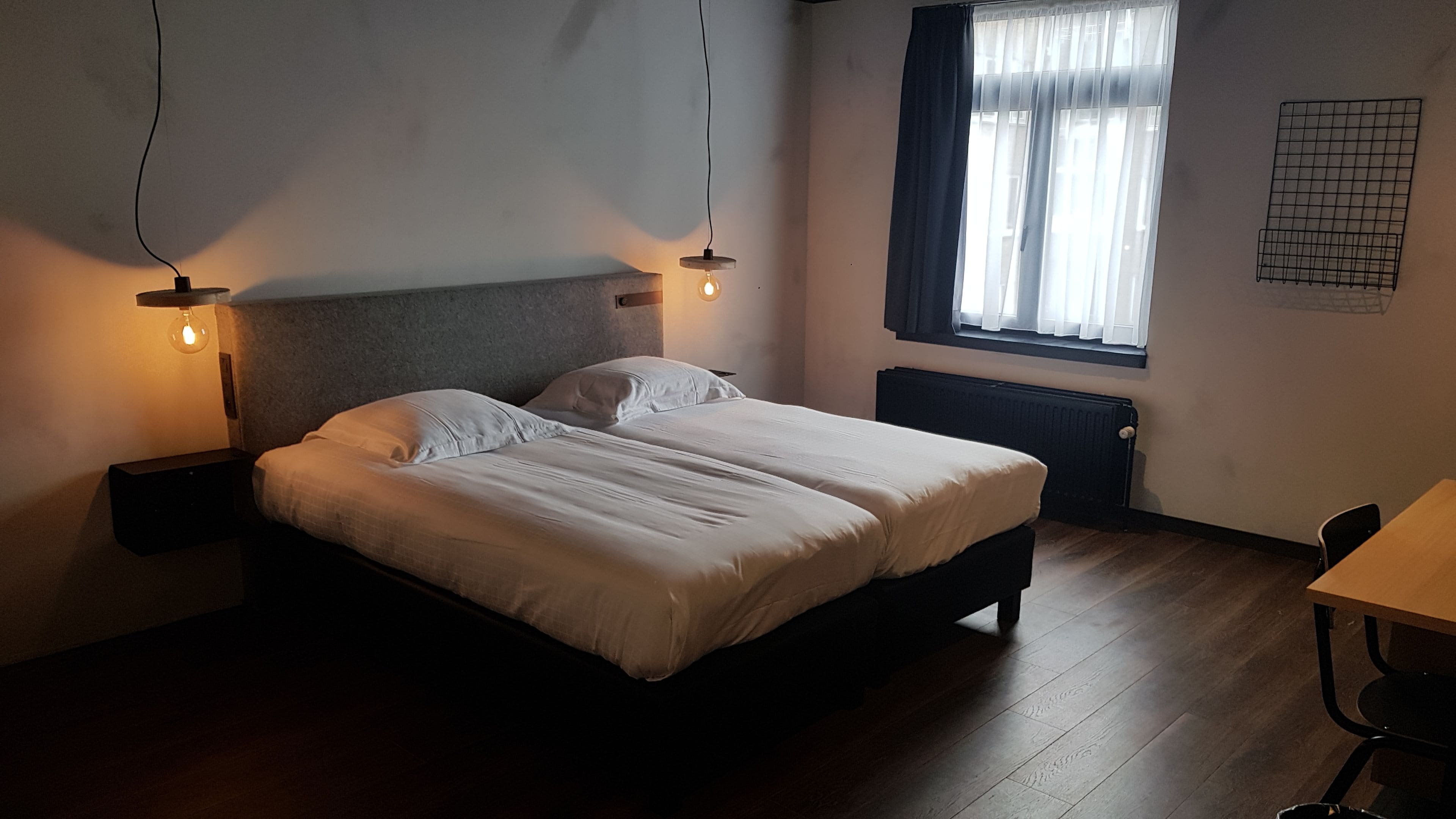 Una cama doble con cabecera, dos mesitas de noche con lámparas, una ventana con cortinas y un radiador.