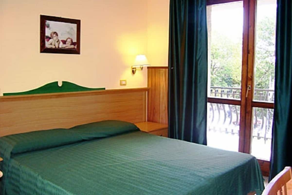 A hotel room with a bed, a wall-mounted picture, a nightstand with a lamp, and a balcony with a railing.