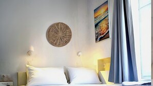 A neatly made bed with white linens, a round woven wall decoration, a painting of a sunset, and a window with curtains. - Tinos Heritage Plateia (Tinos)