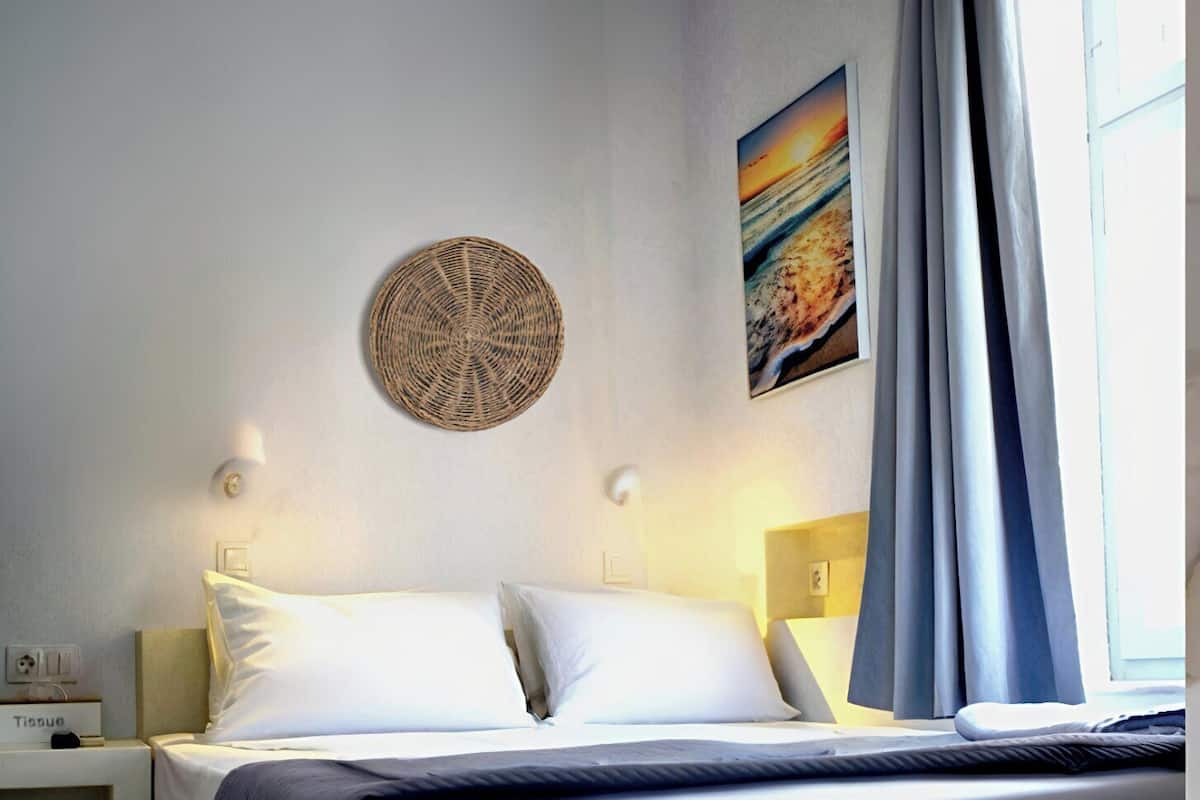 Una cama bien hecha con ropa blanca, un adorno circular tejido en la pared, un cuadro de una puesta de sol y una ventana con cortinas.
