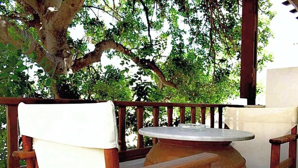 An outdoor seating area with a round table and wooden chairs, surrounded by a wooden railing and a large tree. - Tinos Heritage Plateia (Tinos)