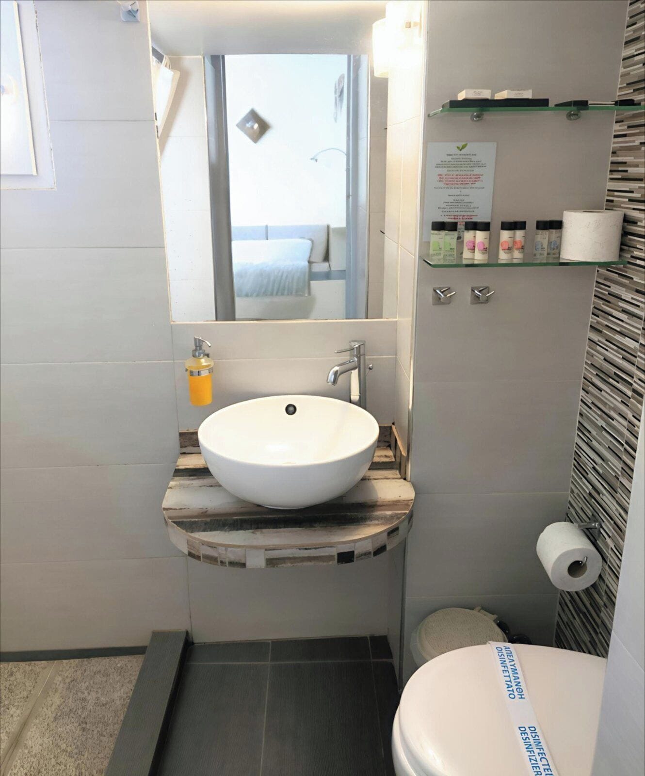 Un baño con lavamanos blanco, un espejo, un inodoro y un estante con artículos de aseo.