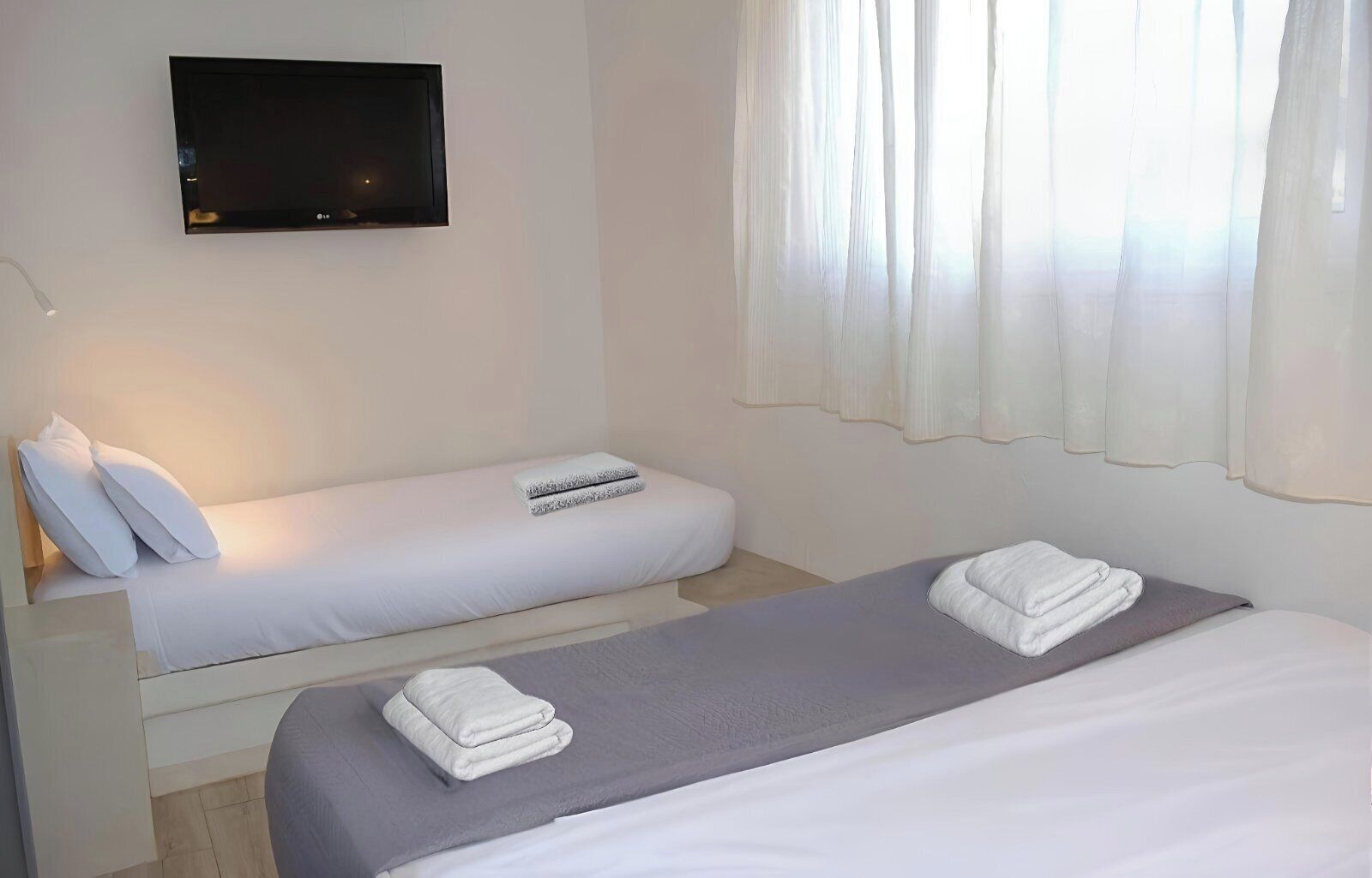 A hotel room with two beds, a flat-screen TV mounted on the wall, and sheer curtains on the window.