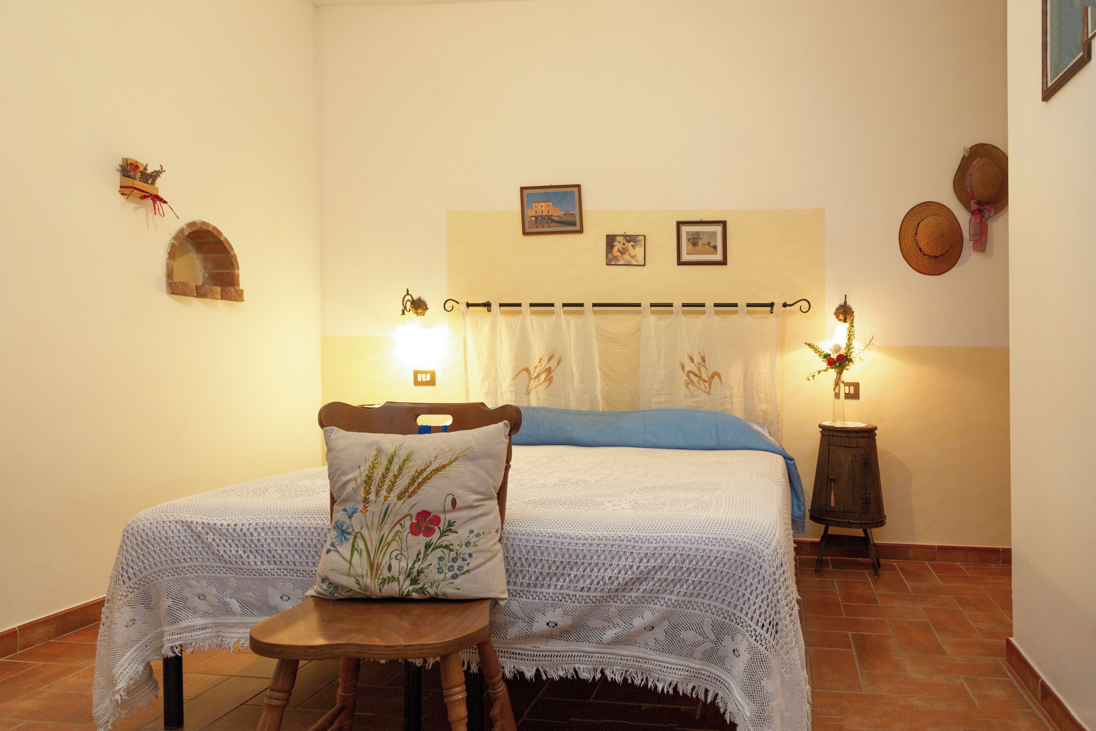Ein gemütliches Schlafzimmer mit einem Bett, einem Sessel und einem kleinen Tisch. An den Wänden befinden sich dekorative Elemente und ein Vorhang mit Blumenmuster.