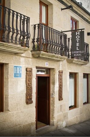 A hotel facade with a wrought iron balcony, a sign reading "HOTEL O PORTELO RURAL," and a blue sign with "HR" indicating a hotel room.