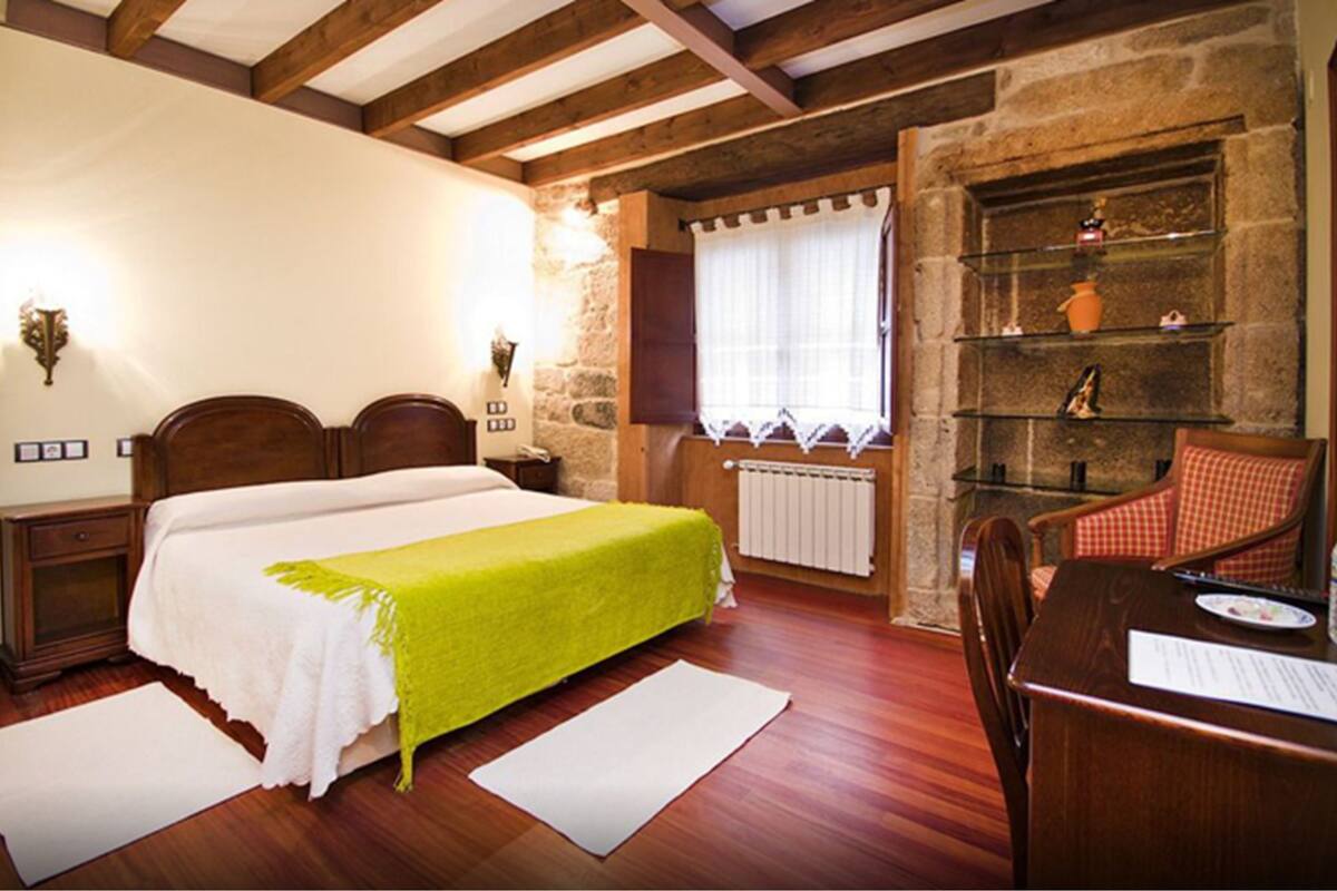 Une pièce comprenant un lit, un bureau, une chaise, un mur en pierre et une fenêtre avec des rideaux.