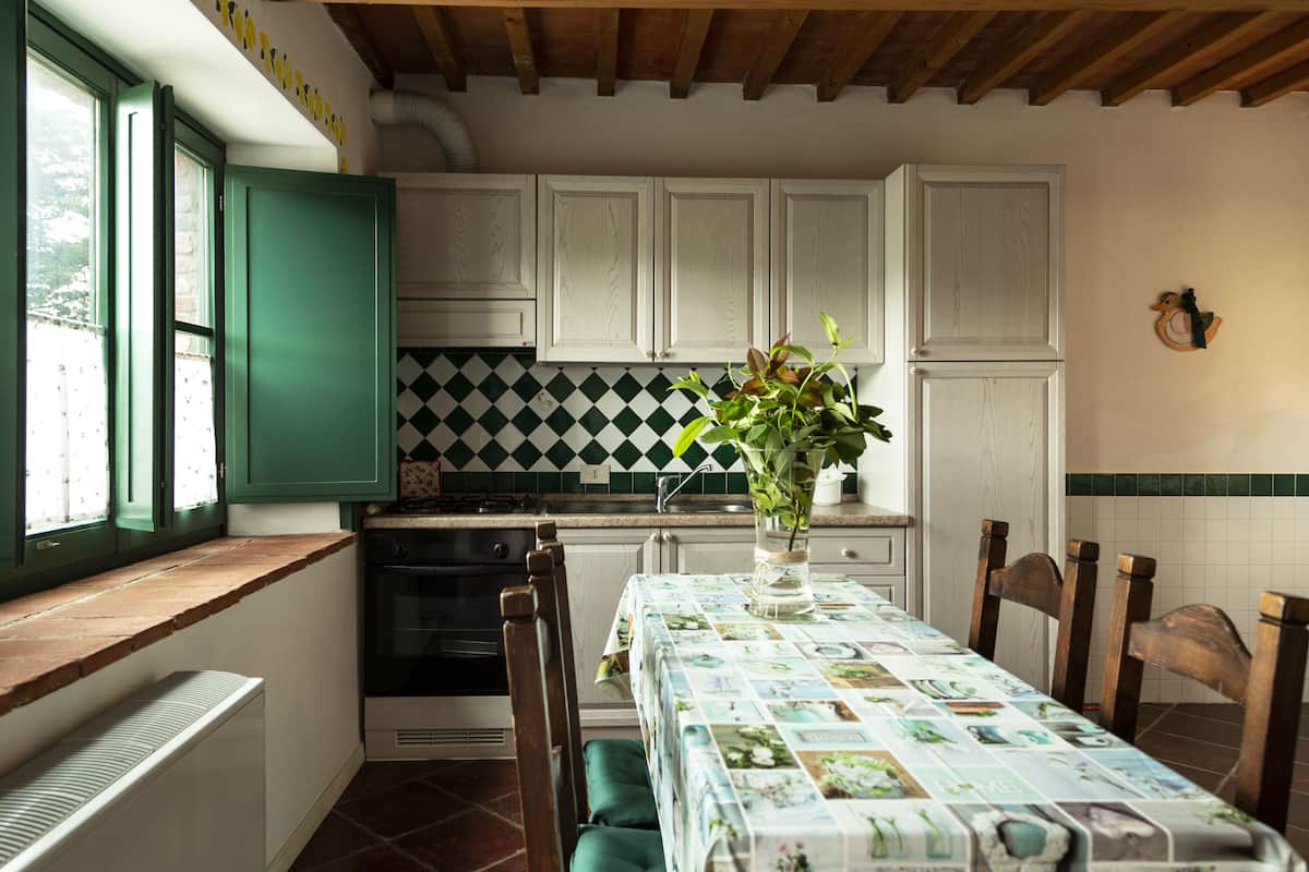Une cuisine avec des meubles blancs, un pare-feu en carreaux, une table à manger recouverte d’une nappe à motifs floraux et une fenêtre avec des volets verts.