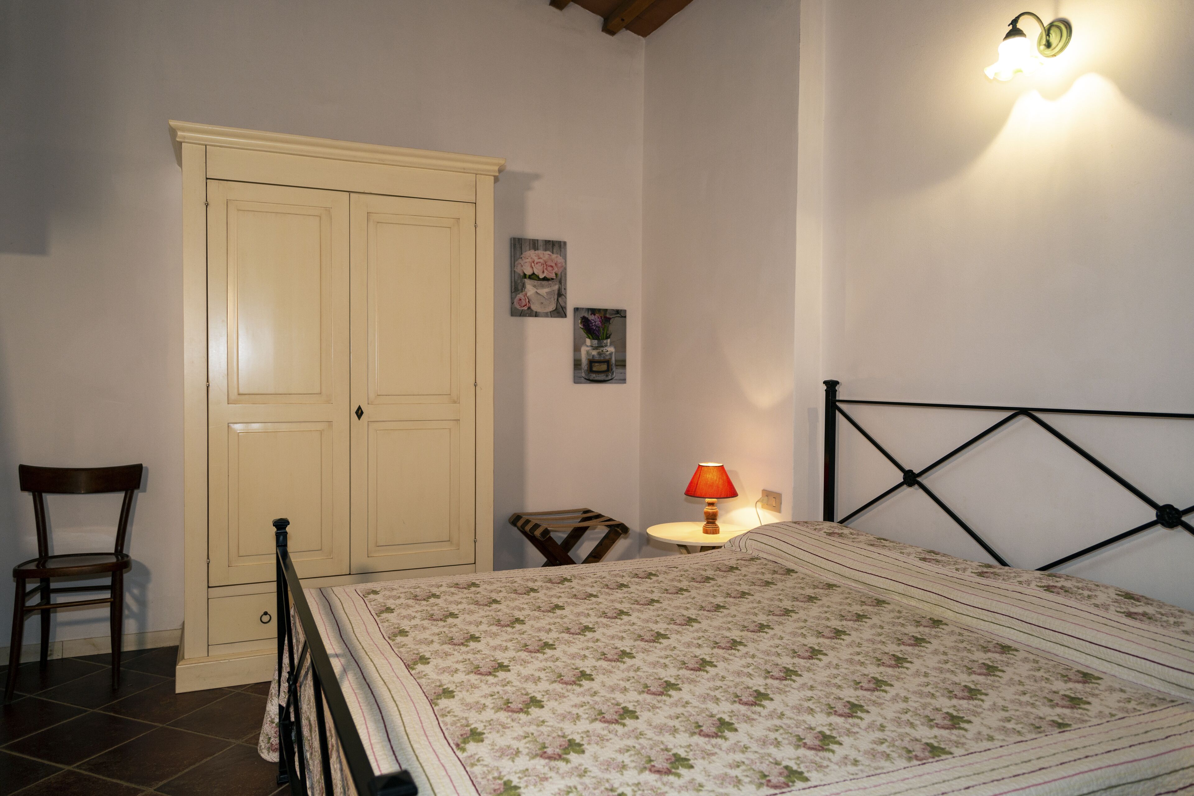 A bedroom with a bed, a wardrobe, a chair, a small table with a lamp, and two wall-mounted pictures.