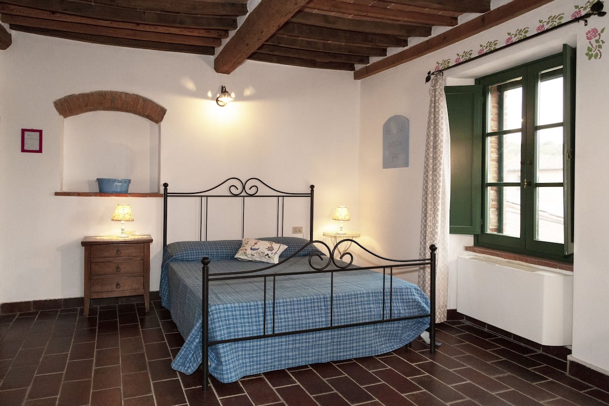 Une chambre avec un lit en métal, une table de chevet en bois, une étagère fixée au mur, une fenêtre avec des volets verts et un sol carrelé.