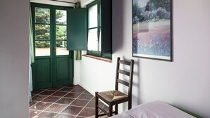 Ein Zimmer mit einem Bett, einem Sessel, einer grünen Tür, einem Fenster mit Blick auf Bäume und einem Gemälde an der Wand.