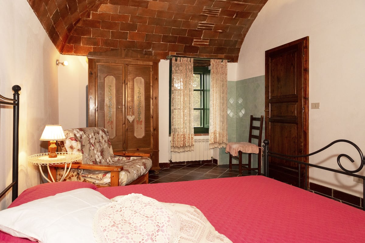Une chambre à coucher avec un lit en bois, une chaise, une table avec une lampe et une fenêtre avec des rideaux.