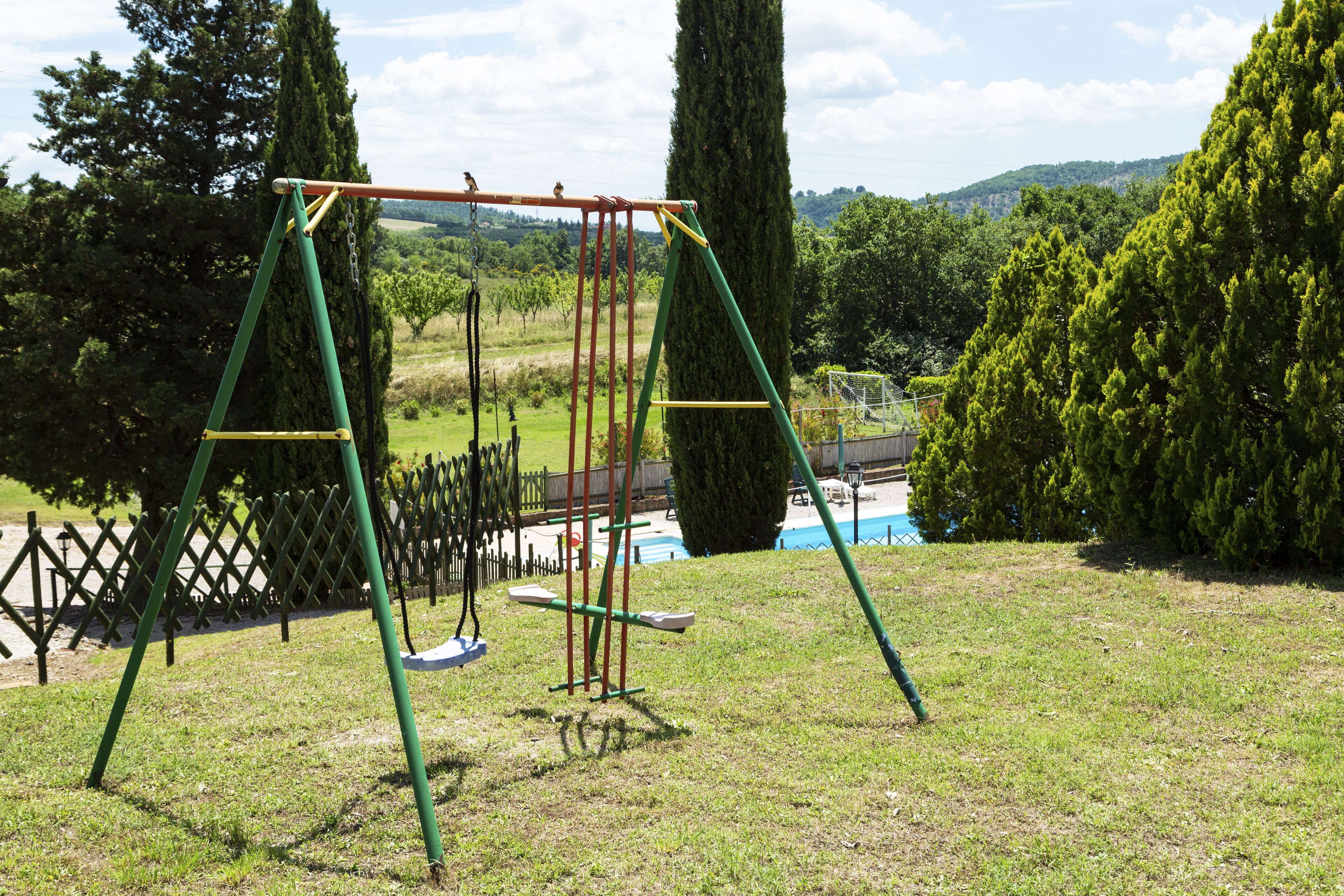 A swing set in a grassy area with a pool and trees in the background.