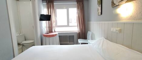 Una habitación de hotel con cama, televisor montado en la pared, ventana con cortinas, radiador y una silla.