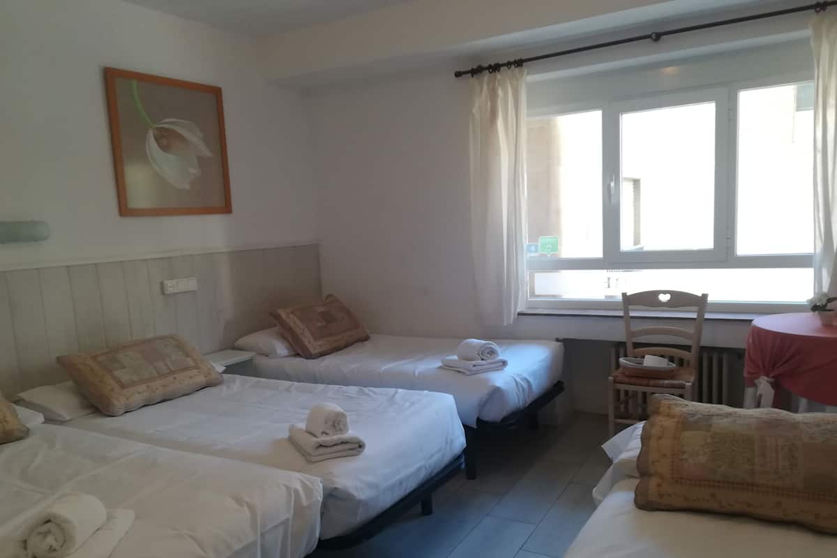 A hotel room with two single beds, a small table, a chair, and a window with curtains.