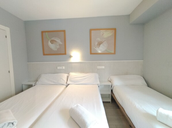 Habitación de hotel con dos camas individuales, una mesita de noche, una lámpara de pared y dos cuadros con motivos florales.