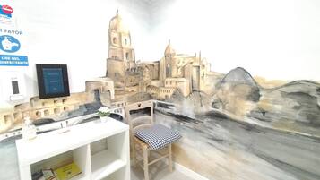 Una habitación con un mural de un castillo, un escritorio con una silla, una estantería con libros y un dispensador de gel hidroalcohólico.