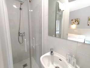 Un baño moderno con ducha de vidrio, un lavamanos blanco con artículos de aseo y un espejo grande que refleja la habitación.