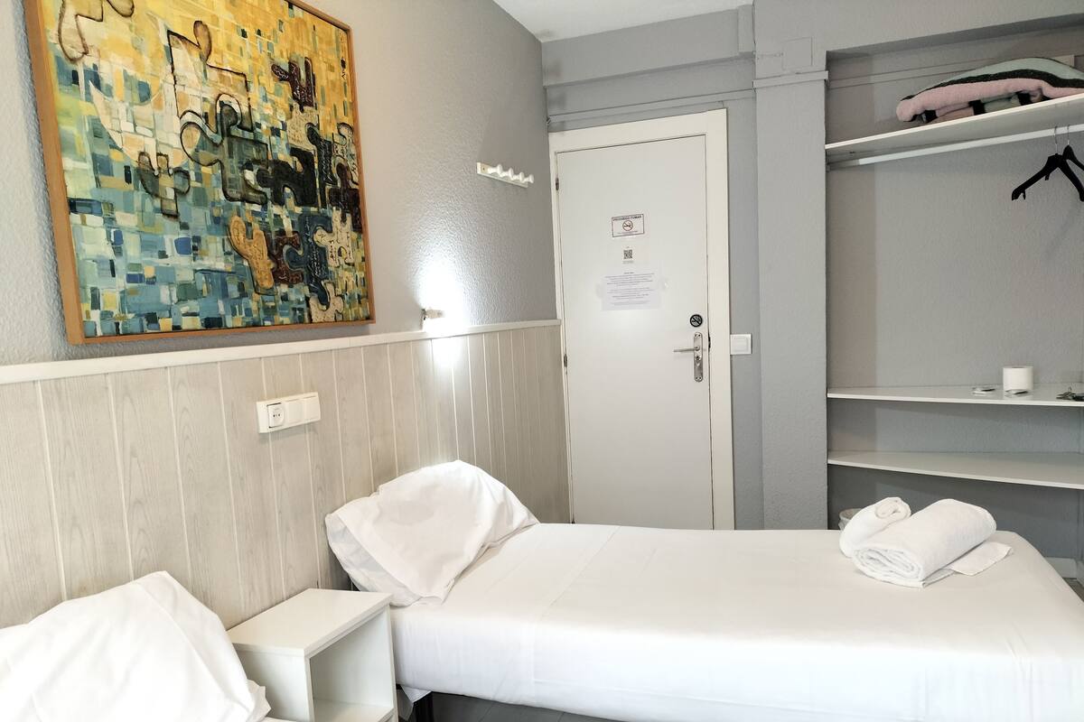 A hotel room with two beds, a painting on the wall, a wardrobe, and a door with a sign.