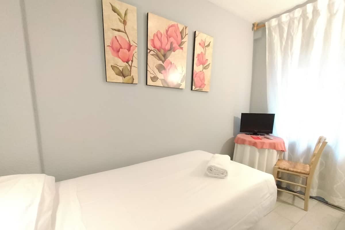 A single bed with a white sheet, a bedside table with a television, and a chair.