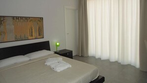 A hotel room with a bed, a nightstand with a lamp, a door, and a large window with curtains. - Dodo House Bed & Breakfast (Nocera Inferiore)