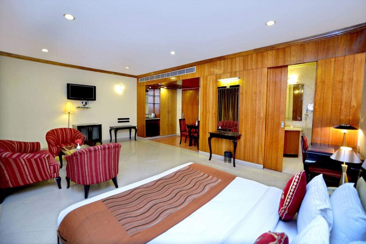Une chambre d’hôtel avec un grand lit, deux fauteuils à rayures rouges, une petite table, un téléviseur et un mur recouvert de panneaux en bois.