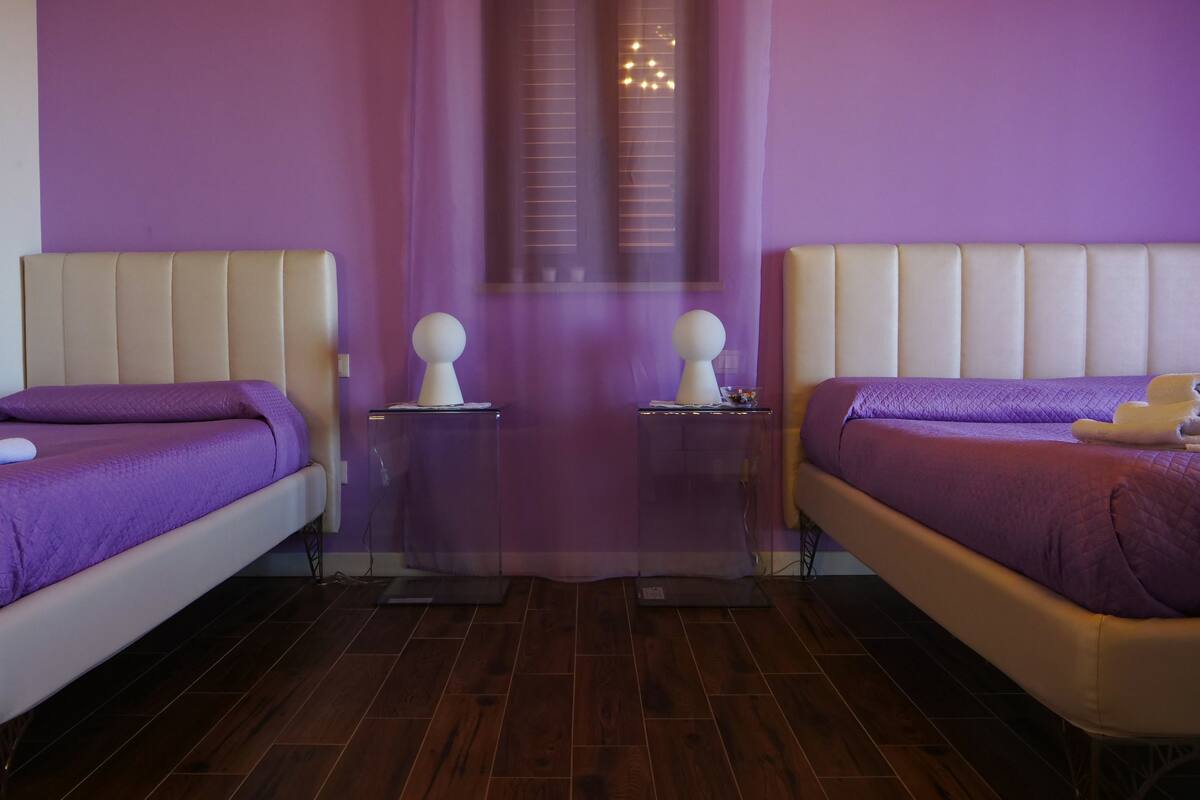 A room with two beds, each with a purple bedspread and white headboard. There are two bedside tables with lamps and a small decorative item on each. The room has wooden flooring and a purple wall.