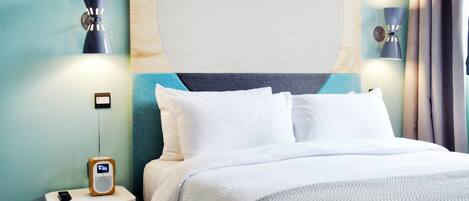 A hotel room with a bed, a bedside table with a phone and a speaker, and wall-mounted lamps.