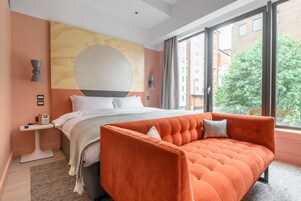 Un hotel moderno con una cama grande, un sofá naranja y vistas de la ciudad a través de amplios ventanales.