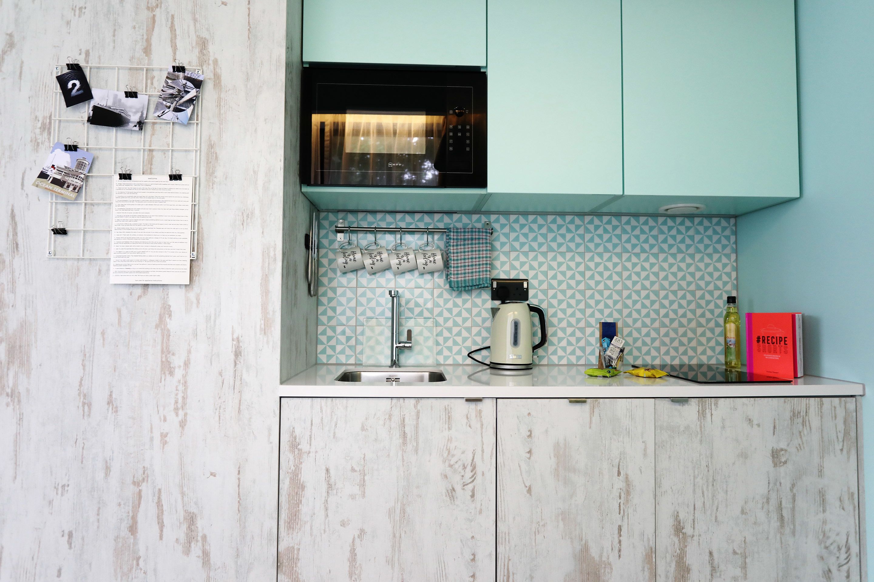 A kitchen with teal cabinets, a white countertop, a kettle, a coffee maker, and a red cookbook.