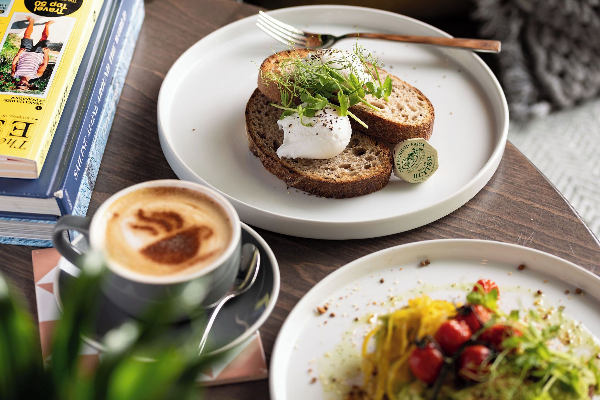 Une table à manger avec une assiette de bruschettas garnies de mozzarella et de verdure, une tasse de café et un livre.
