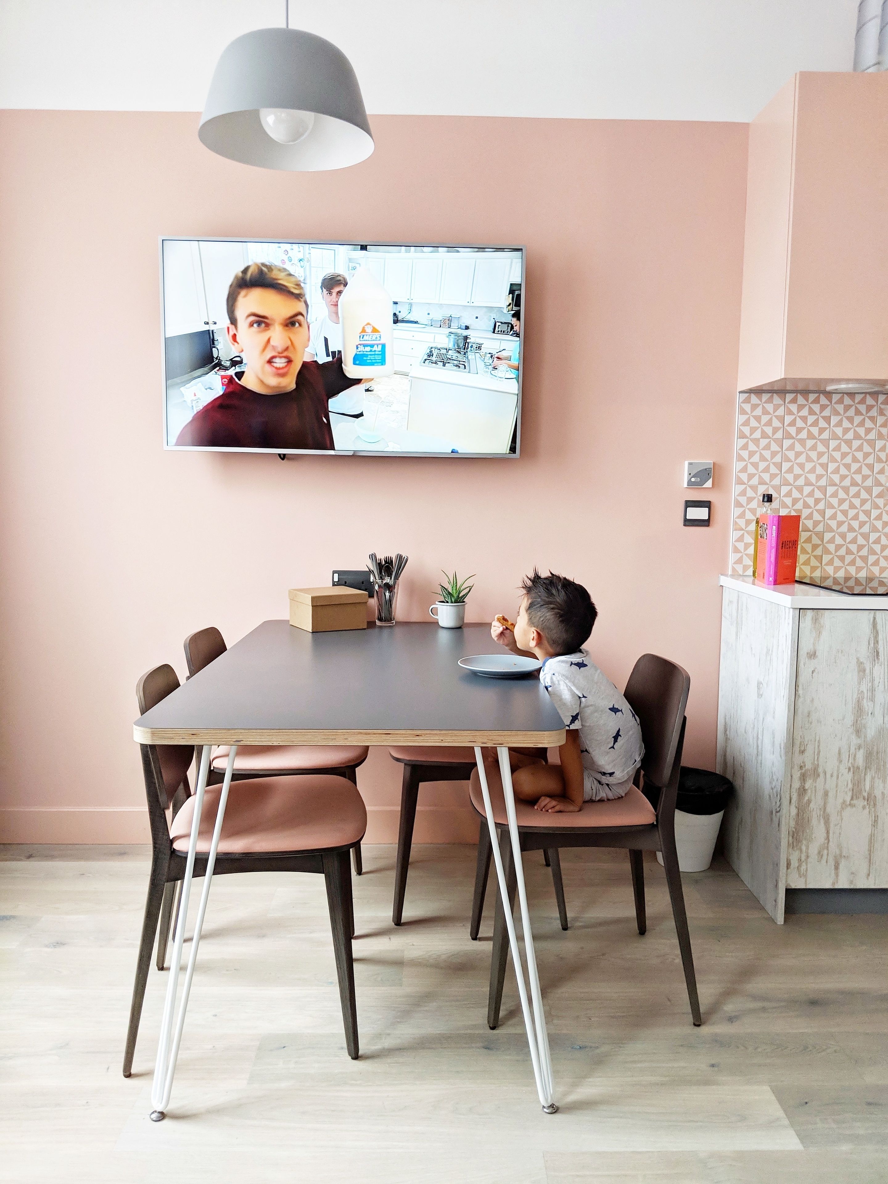 Un coin repas avec une table, des chaises, un téléviseur sur lequel on voit une personne, et un enfant qui mange.